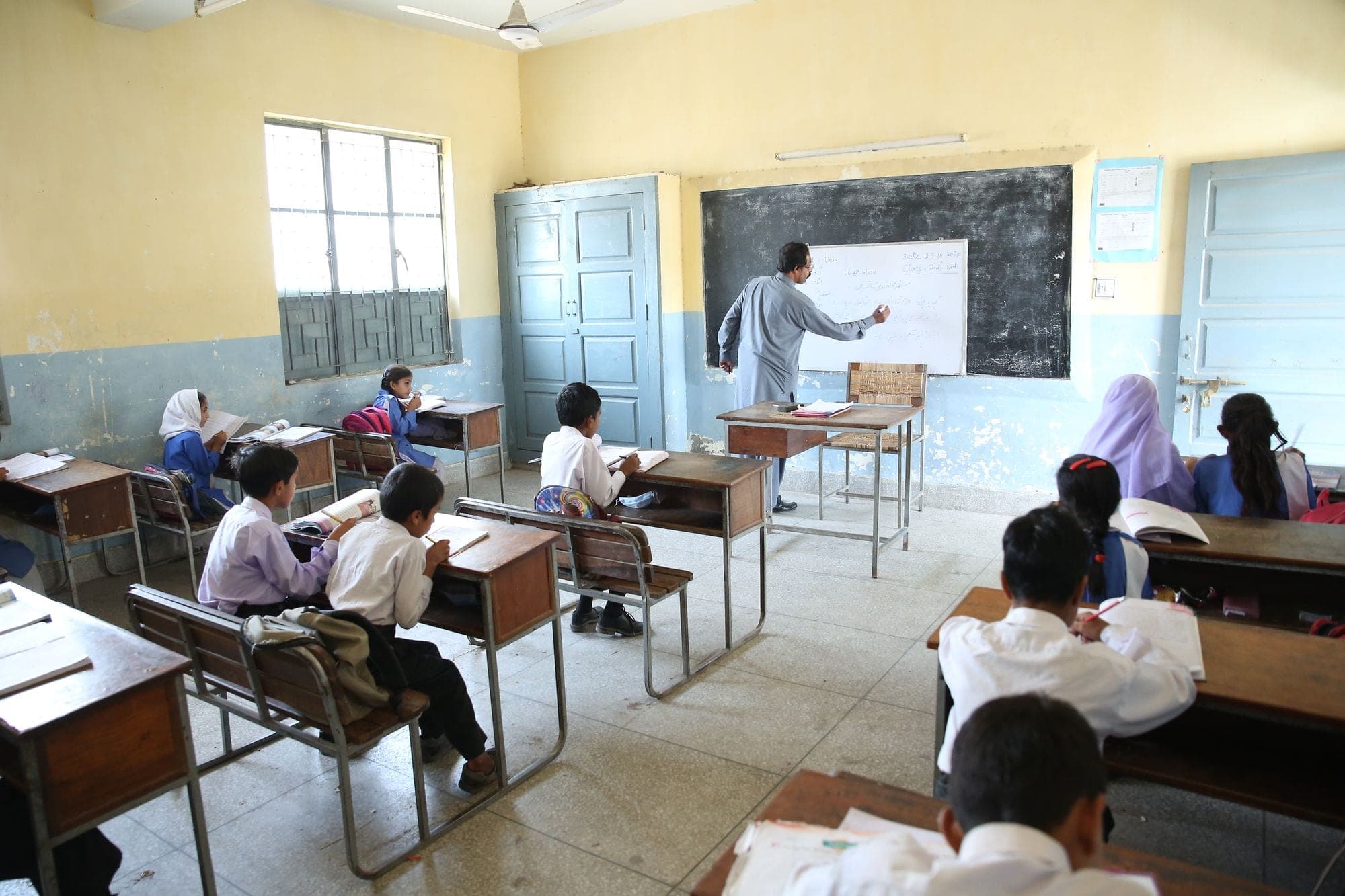 Students at wooden desks listening to a teacher writing on the board